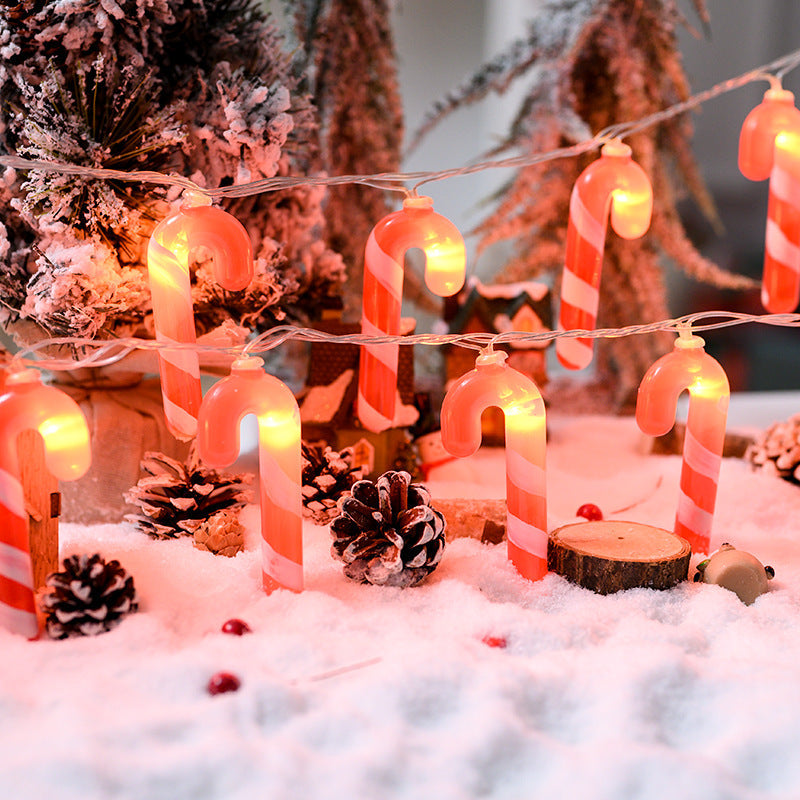 Decorative string lights shaped like candy canes and candles on a snowy surface with Christmas trees in the background.